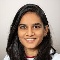 Dr. Desai has long dark hair, is wearing a white medical coat, and is smiling at the camera