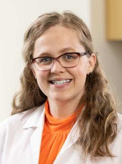 Dr. Blumrick has long wavy blonde hair, is wearing glasses and an orange blouse under a white medical coat, and is standing in a clinic with arms crossed while smiling at the camera